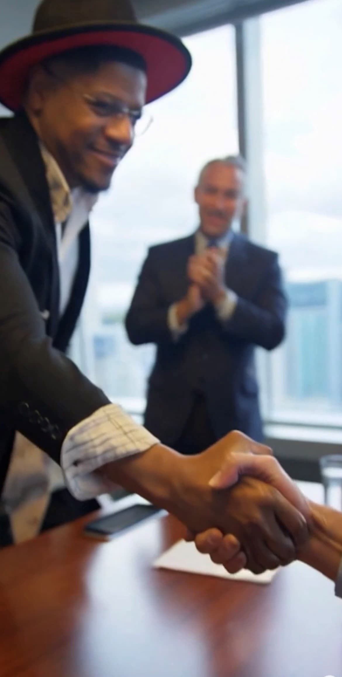 A smiling man in a hat shakes hands with a colleague while another man applauds.