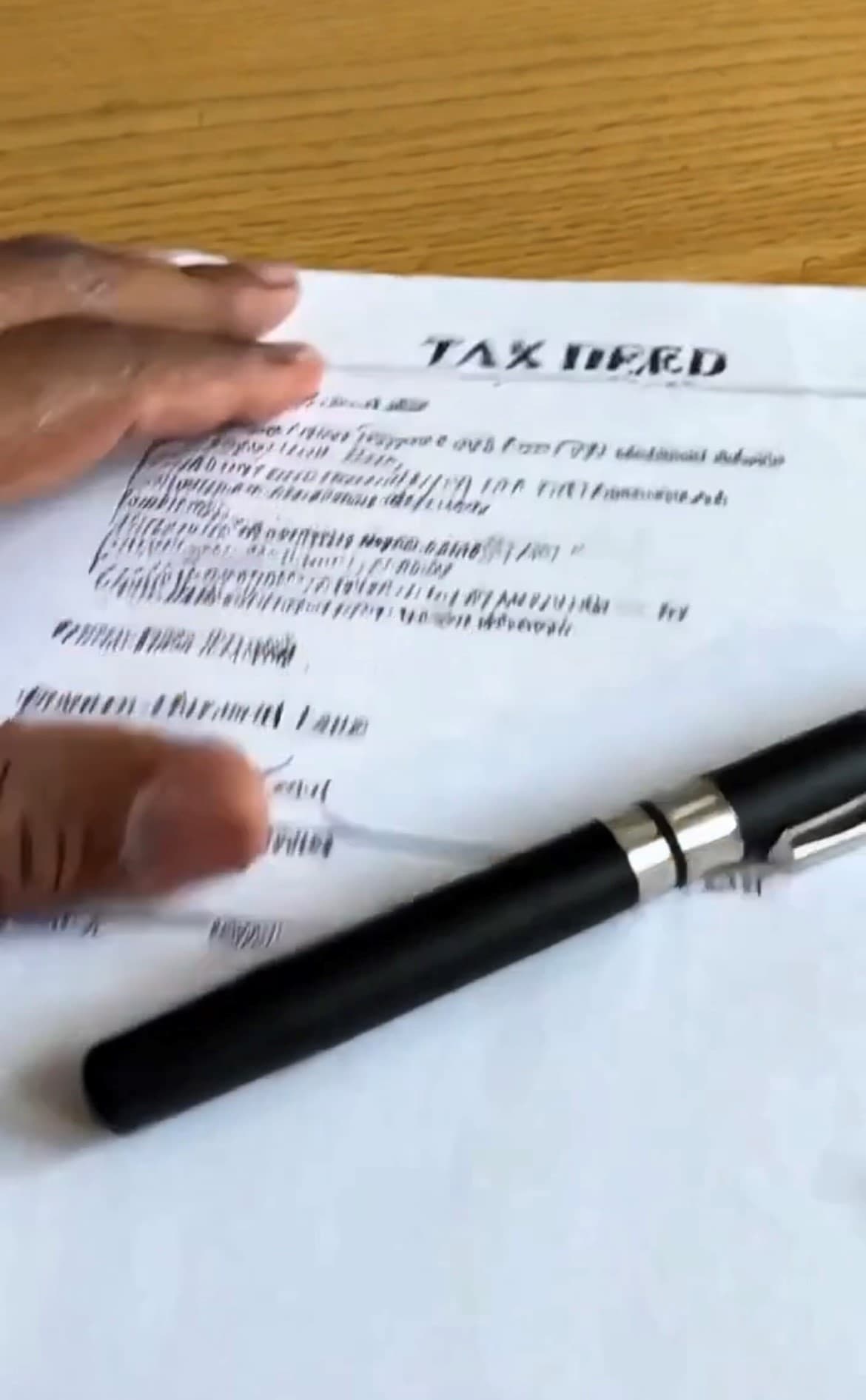 Hand holding a TAX DEED document on a wooden table with a black pen.